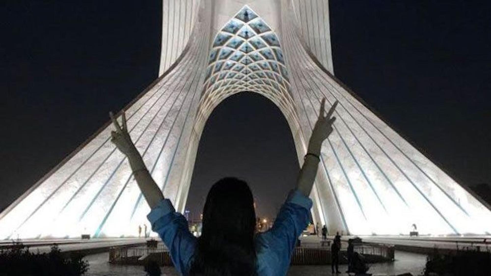 Eine junge Frau steht ohne das vorgeschriebene Kopftuch vor dem Azadi-Turm (Freiheitsturm) und zeigt mit beiden Händen das Sieges-Zeichen.