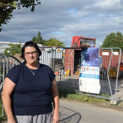 Eine Frau im blauen T-Shirt steht vor dem Grundstück, auf dem Baumaschinen geparkt werden. Im Hintergrund stehen Container und ein Bauwagen.