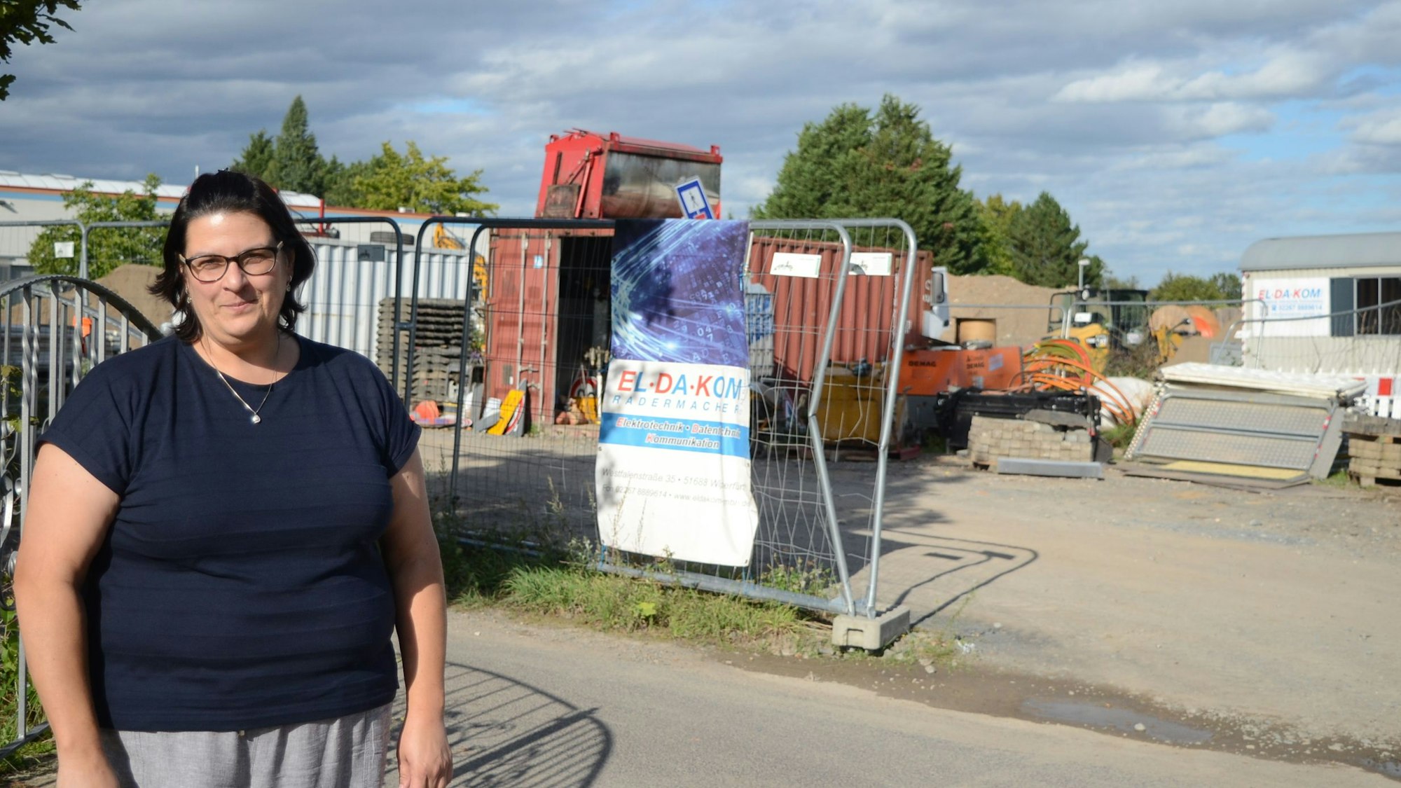 Eine Frau im blauen T-Shirt steht vor dem Grundstück, auf dem Baumaschinen geparkt werden. Im Hintergrund stehen Container und ein Bauwagen.