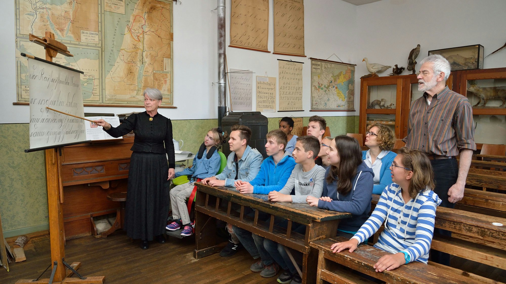 Jugendliche sitzen auf historischen Schulbänken aus Holz. An der Tafel steht eine als Lehrerin in der früheren Zeit in einem schwarzen langen Kleid verkleidete Mitarbeiterin des Museums.