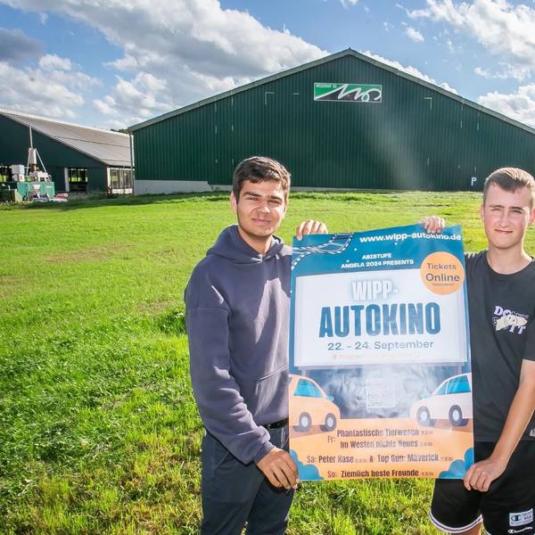 Zwei junge Männer stehen auf einer Wiese und halten ein Veranstaltungsplakat für das erste Wipperfürther Autokino in den Händen. Im Hintergrund ist eine große Scheune eines Landwirtschaftsbetriebs zu sehen.