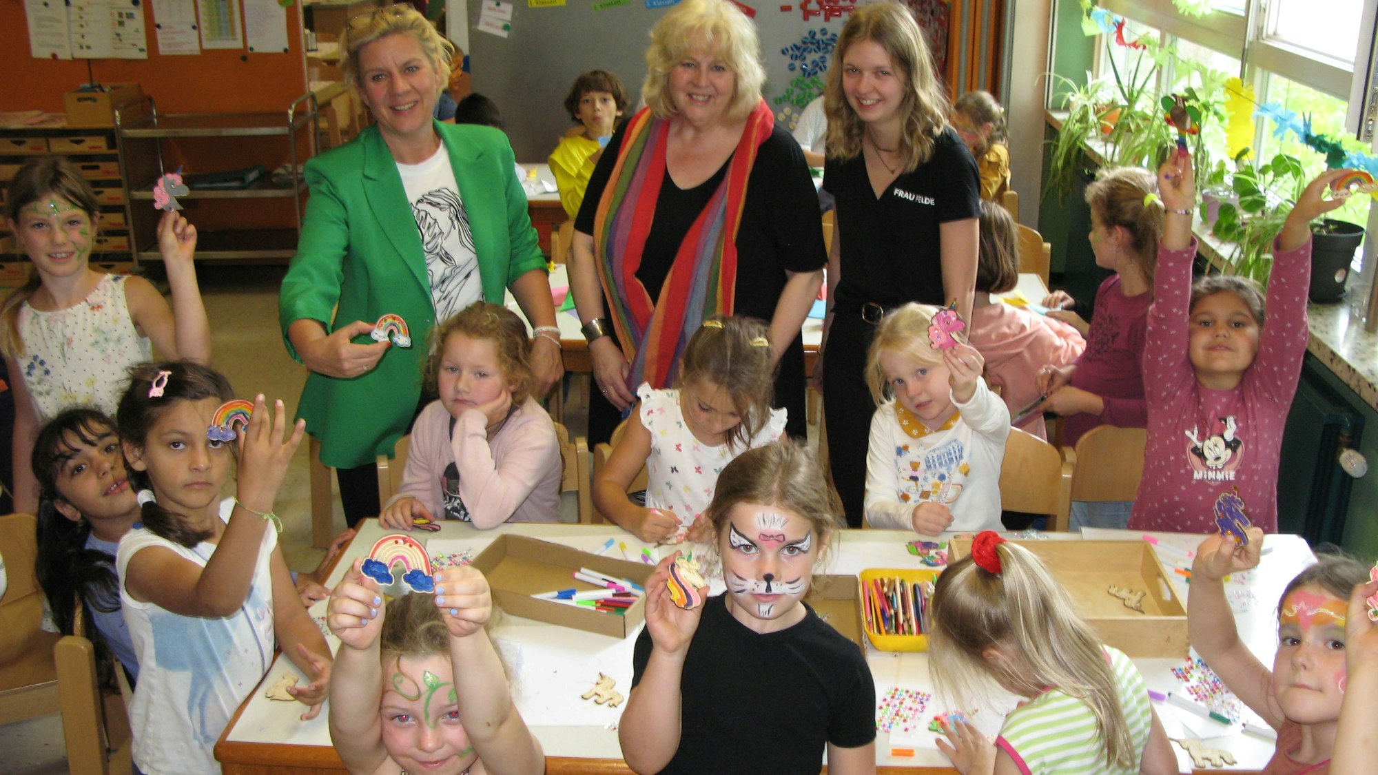 Elf Mädchen der Grundschule malen und basteln an einem Tisch. Dahinter stehen Schulleiterin Monic Beck, OGS-Leiterin Beate Schmitt-Welsch und OGS-Betreuerin Lea Felder.