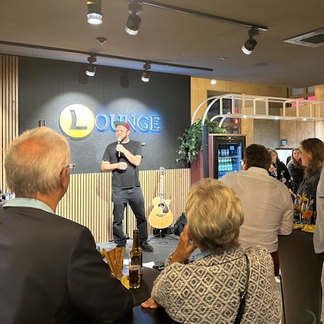 Ein Mann steht auf der Bühne, mehrere Leute schauen ihm zu.