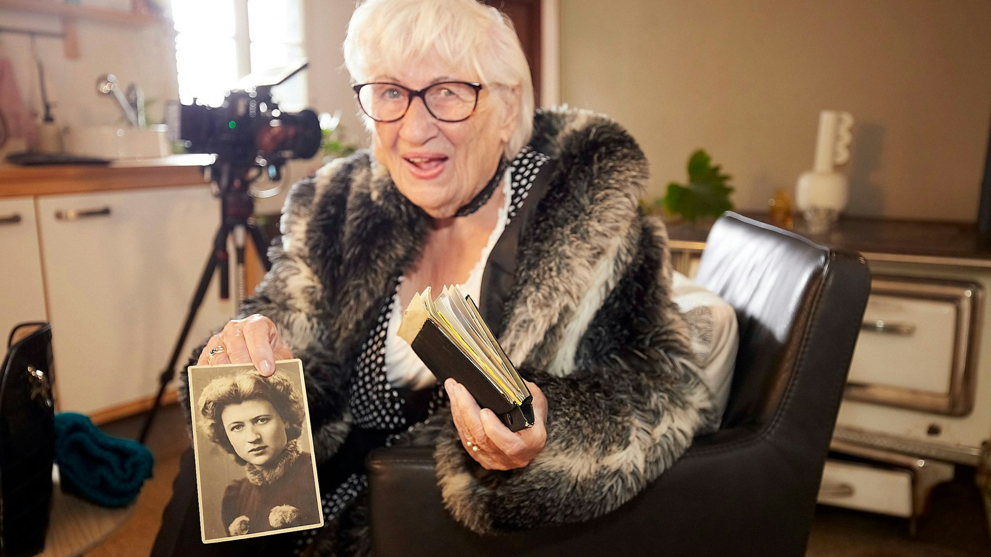 Die knapp 100 Jahre alte Liesel Buchheldt sitzt in einem Sessel. Sie hält eine Mappe in der einen Hand und zeigt mit der anderen ein Schwarz-Weiß-Foto in die Kamera, das sie als junge Frau zeigt.