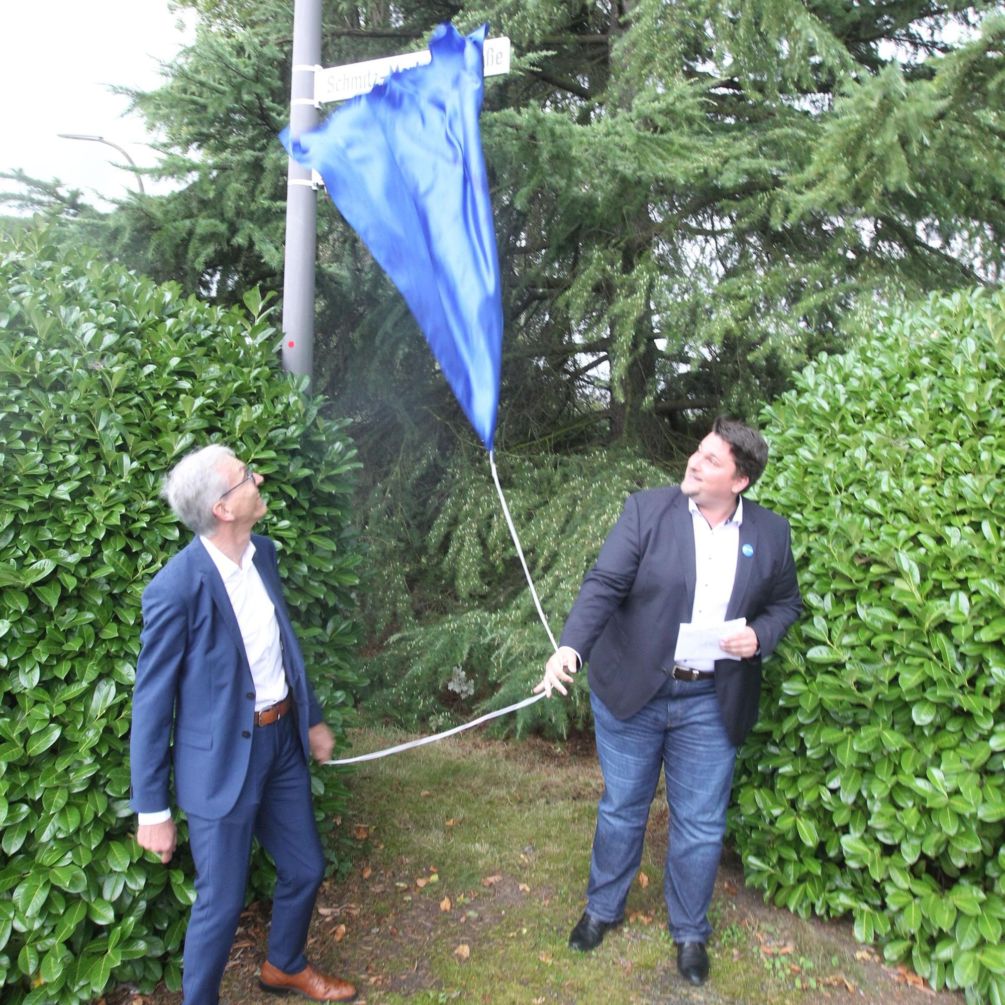 Bürgermeister Alexander Biber und Wolfgang Schmitz-Mertens (links) enthüllen das neue Straßenschild.