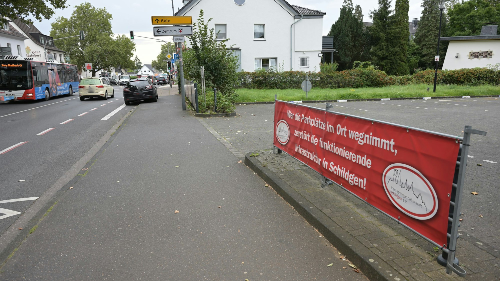Am Straßenrand ist an einer Halterung der Protestbanner befestigt, der Hintergrund ist rot. Mit weißen Druckbuchstaben ist zu lesen: "Wer die Parkplätze wegnimmt, zerstört die funktionierende Infrastruktur in Schildgen!"