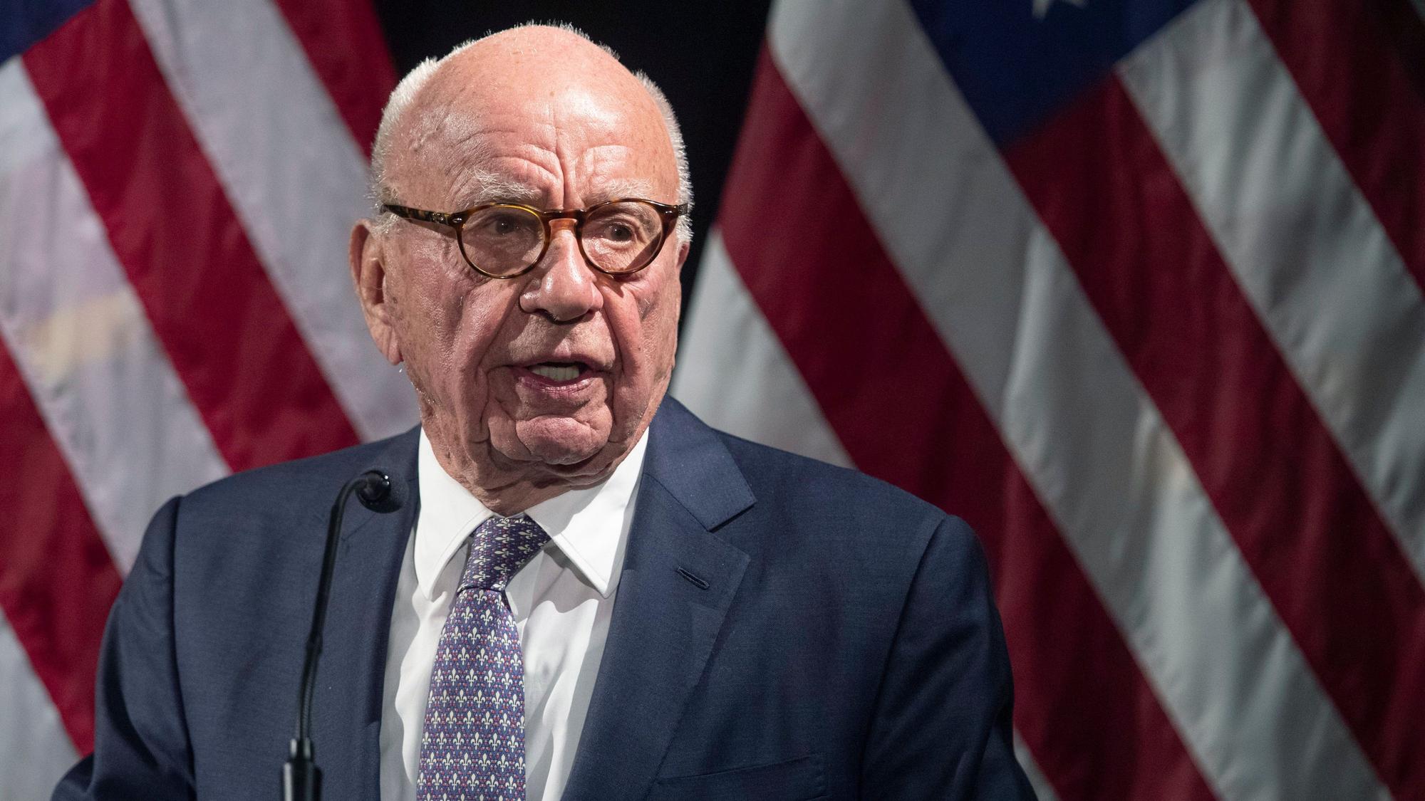 Rupert Murdoch steht im Anzug vor unscharfen amerikanischen Flaggen im Hintergrund und spricht.