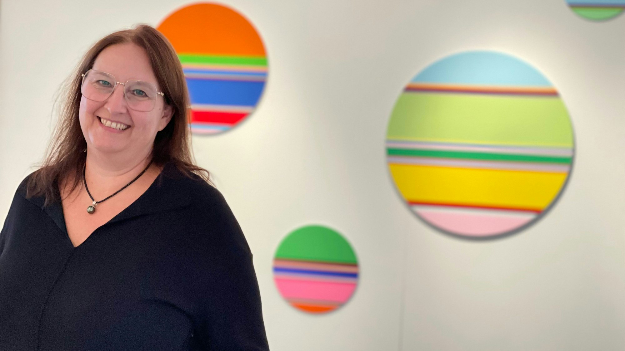 Eine lächelnde, schwarz gekleidete Frau steht vor runden Bildern, auf denen in knalligen Farben akkurate Streifen gemalt sind.