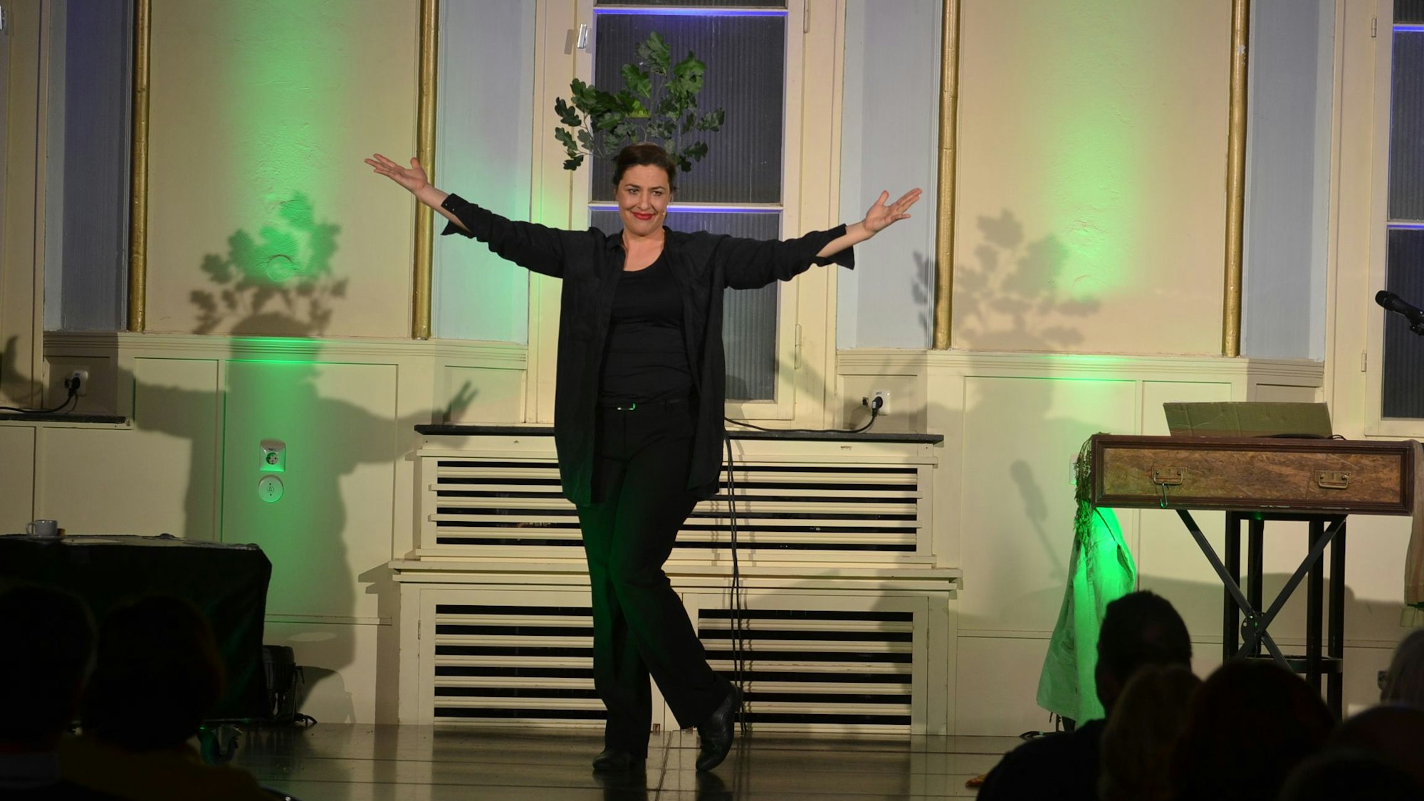 Die Kabarettistin steht in Schwarz gekleidet auf einer Bühne. Sie hat beide Arme ausgebreitet und lächelt.