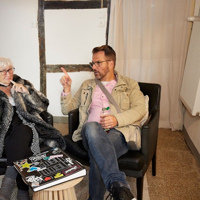 Ingo Wolfgarten und seine Großmutter Liesel Buchheldt sitzen in schwarzen Sesseln und unterhalten sich. Vor ihnen auf dem Tisch liegt ein Fotoalbum.