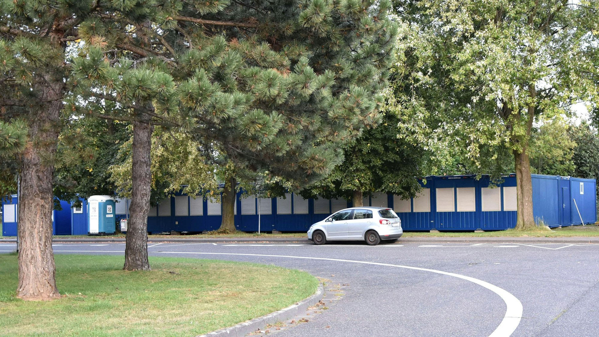 Unter alten Bäumen stehen blaue Container, davor parkt ein Auto.