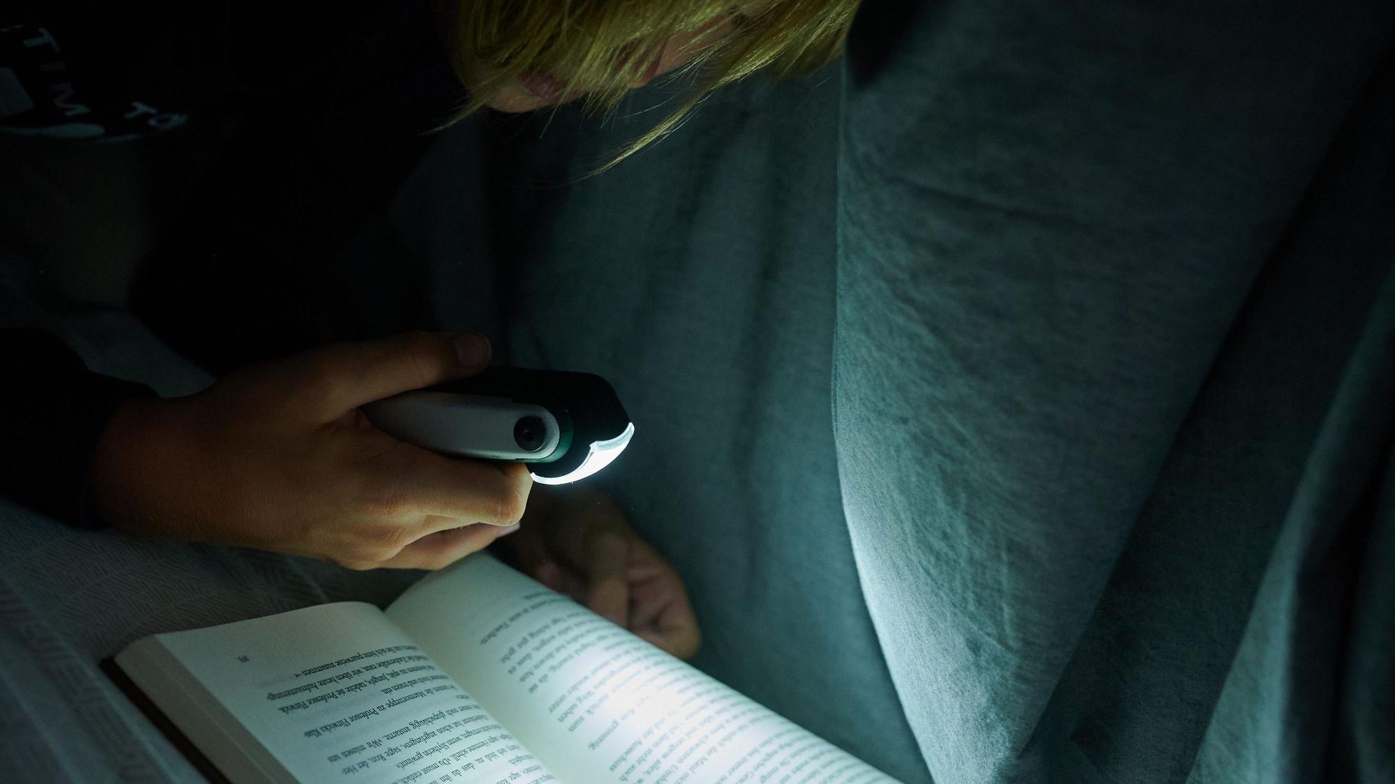 Ein Kind liegt im dunklen Zimmer auf dem Bett und liest mit einer kleinen Leuchte ein Buch. (gestellte Szene)