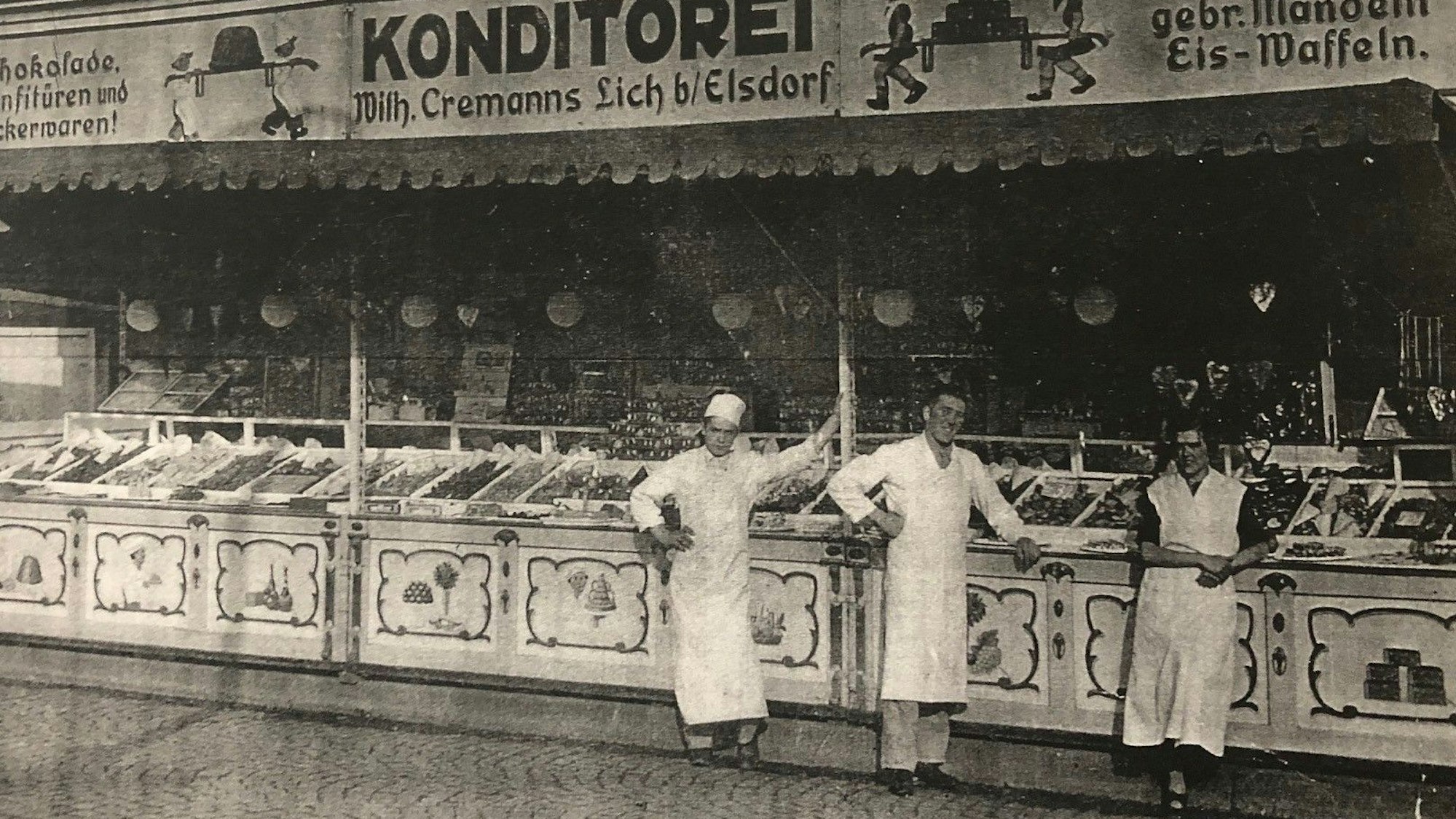 Ein altes Schwarz-Weiß-Foto von einem Süßwarenstand, vor dem Menschen in weißen Schürzen stehen.