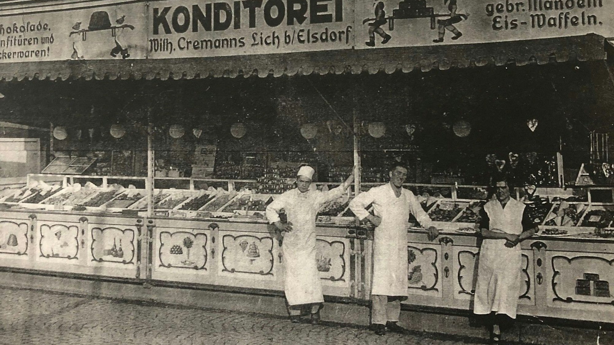 Ein altes Schwarz-Weiß-Foto von einem Süßwarenstand, vor dem Menschen in weißen Schürzen stehen.