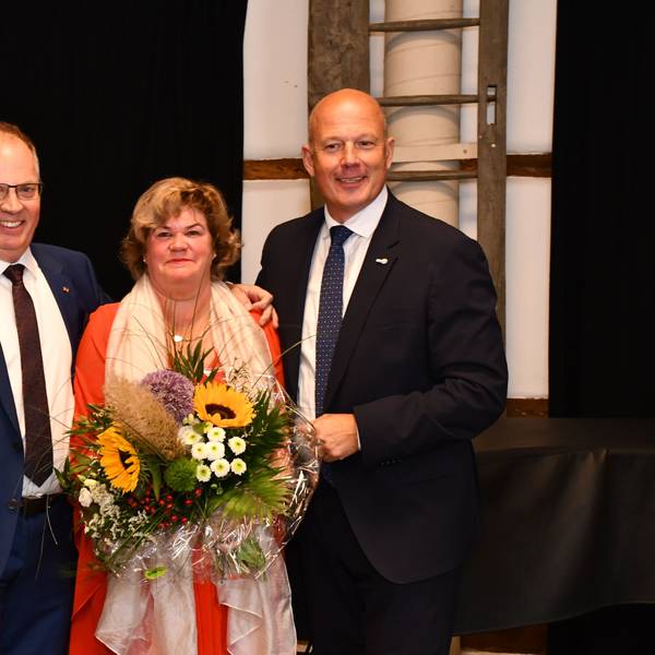 Ein Mann mit Orden am Revers, eine Frau mit Blumenstraße, und ein Mann um dunklen Anzug stehen auf einer Bühne.