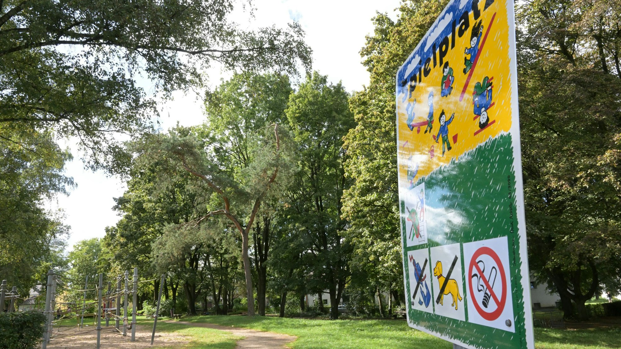 Ein Spielplatz-Schild steht an einem Spielplatz mit Klettergerüsten, Bäumen und Wiese.