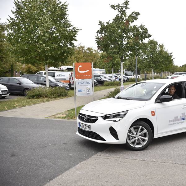 Ein Mann fährt mit einem weißen Auto auf einem Parkplatz. Im Hintergrund sind Bäume und geparkte Autos zu erkennen.
