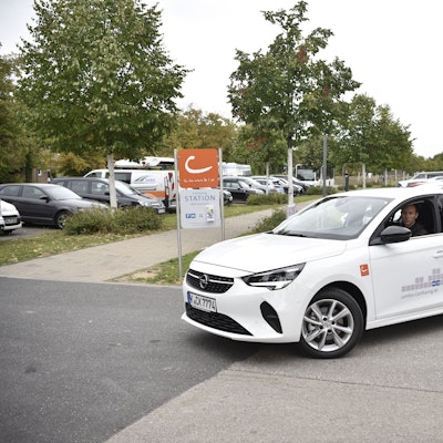 Ein Mann fährt mit einem weißen Auto auf einem Parkplatz. Im Hintergrund sind Bäume und geparkte Autos zu erkennen.
