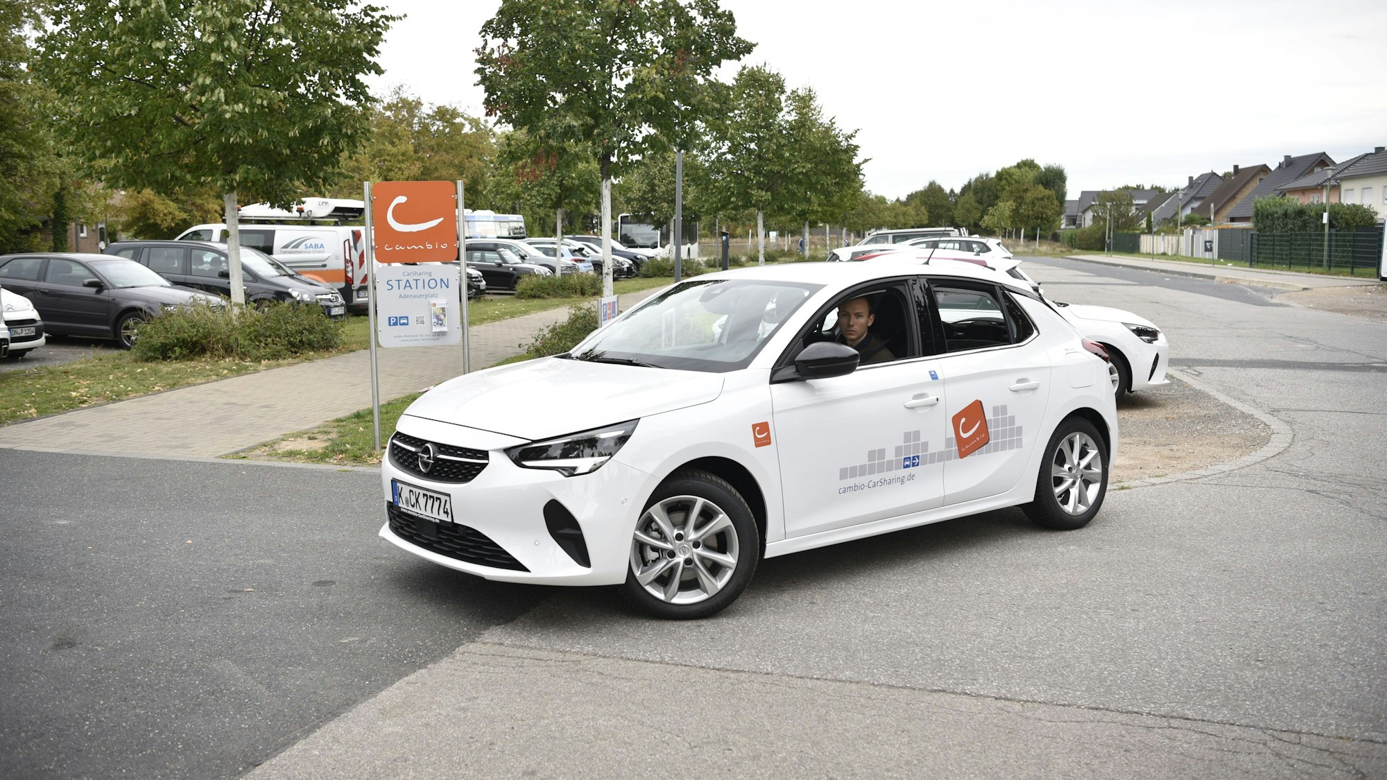 Ein Mann fährt mit einem weißen Auto auf einem Parkplatz. Im Hintergrund sind Bäume und geparkte Autos zu erkennen.