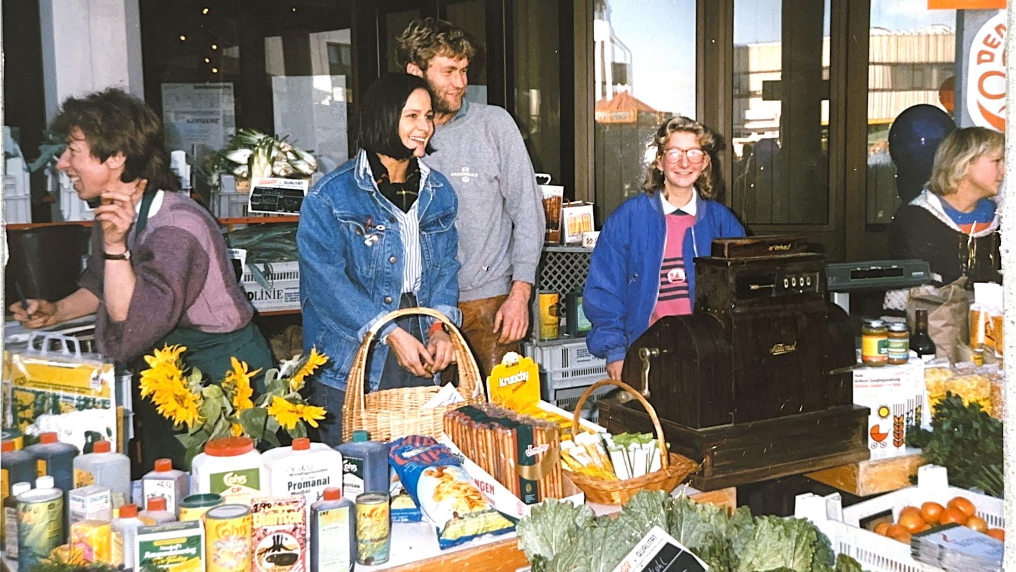 Ein altes Foto, auf dem hinter einem Marktstand mit Dosen, Flaschen und Frischgemüse ein Mann und eine Frau stehen.