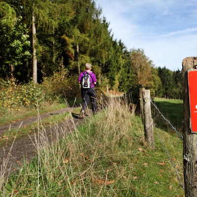 Eine Wanderin geht auf einem Weg, an dem die Wegemarkierung des Fachwerkwegs mit einer weißen 22 auf rotem Grund montiert ist.