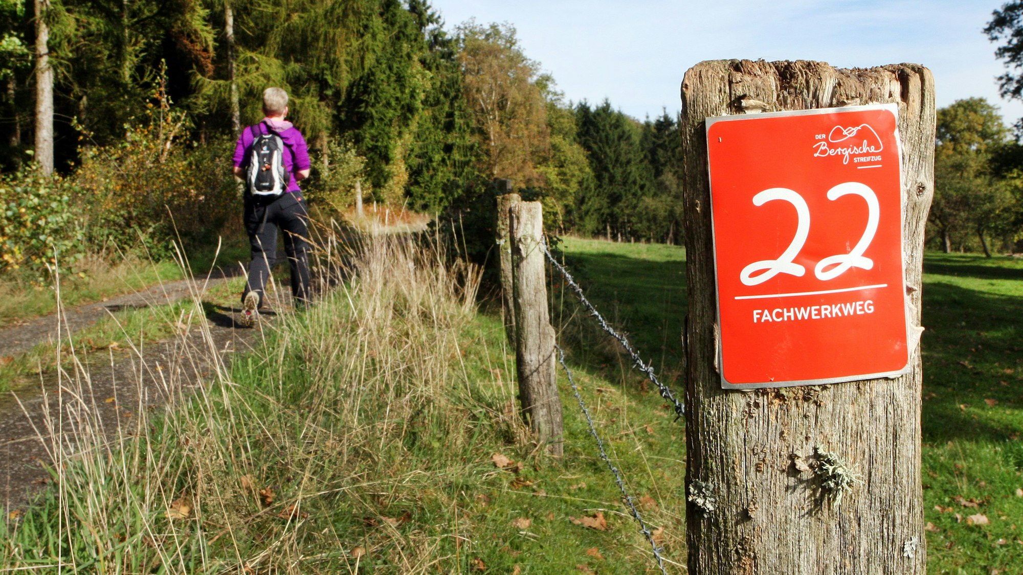 Eine Wanderin geht auf einem Weg, an dem die Wegemarkierung des Fachwerkwegs mit einer weißen 22 auf rotem Grund montiert ist.