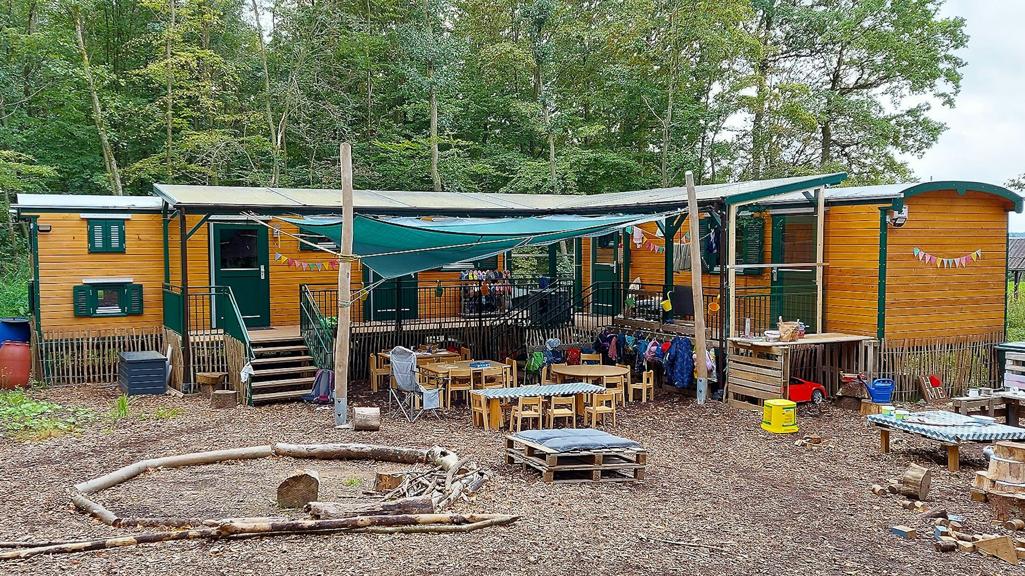 In Bauwagen aus Holz ist der Waldkindergarten Mülheim eingerichtet. Im Vordergrund sind Tische, Stühle und Spielsachen zu sehen, im Hintergrund Bäume.