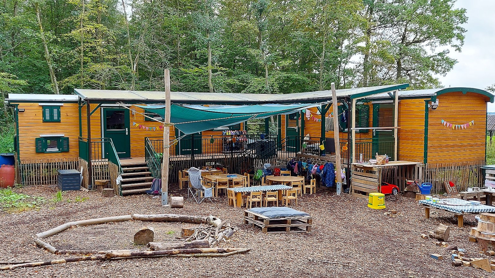 In Bauwagen aus Holz ist der Waldkindergarten Mülheim eingerichtet. Im Vordergrund sind Tische, Stühle und Spielsachen zu sehen, im Hintergrund Bäume.