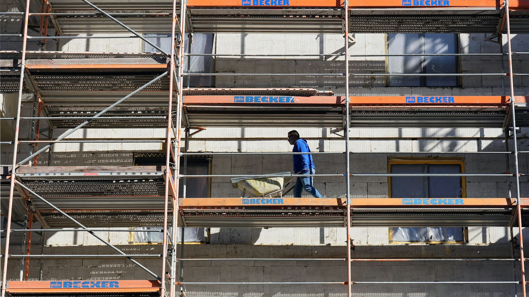 Ein Arbeiter geht auf der Baustelle eines Wohnhauses am Franz-Mehring-Platz mit einer Schubkarre über das Baugerüst.