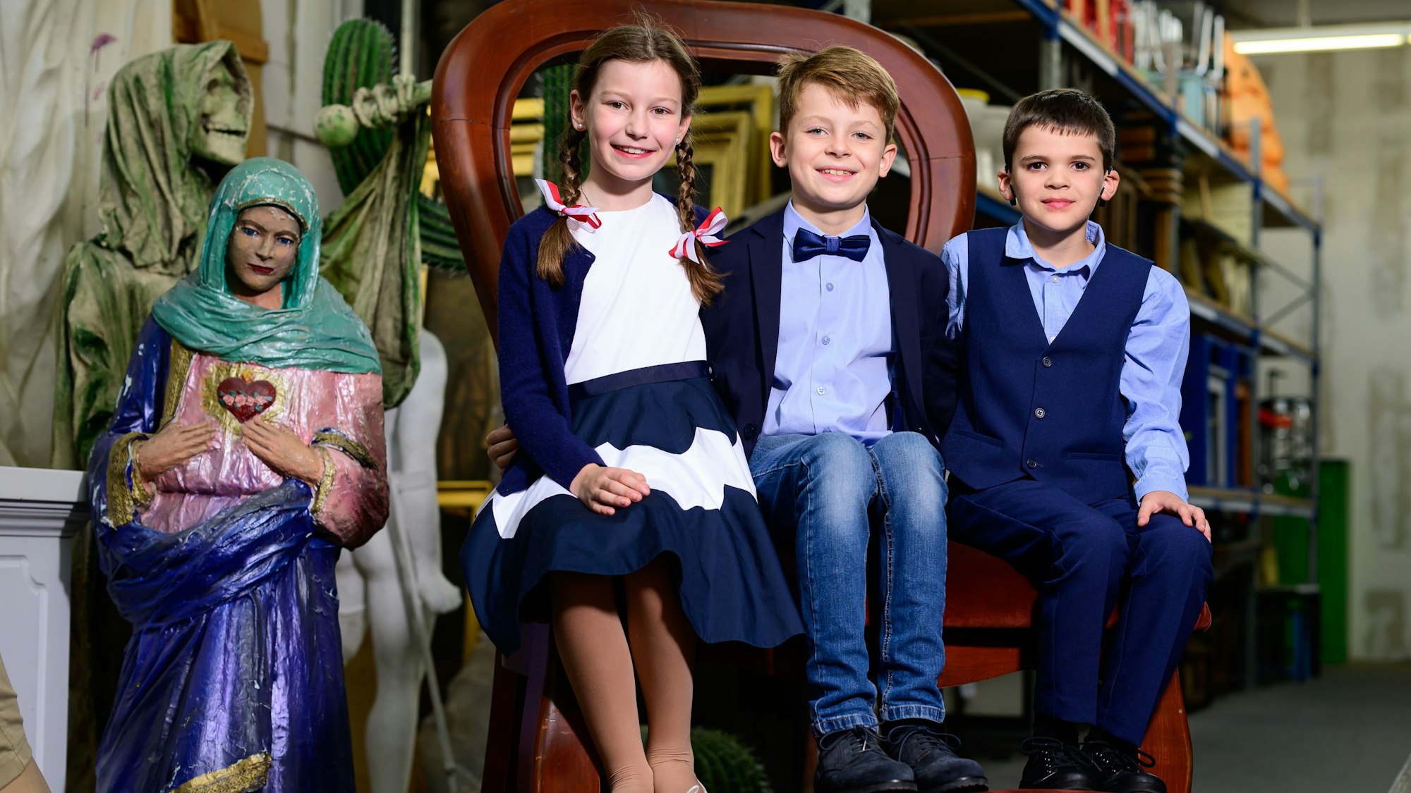 Kleine Jecke auf riesigem Stuhl: Jungfrau Benediktavon Stülpnagel (v.l.), Prinz Julius Kürten und Bauer Severin Rombach inmitten der Requisiten der Kölner Bühnen. Sie sind das designierte Kölner Kinderdreigestirn 2024.