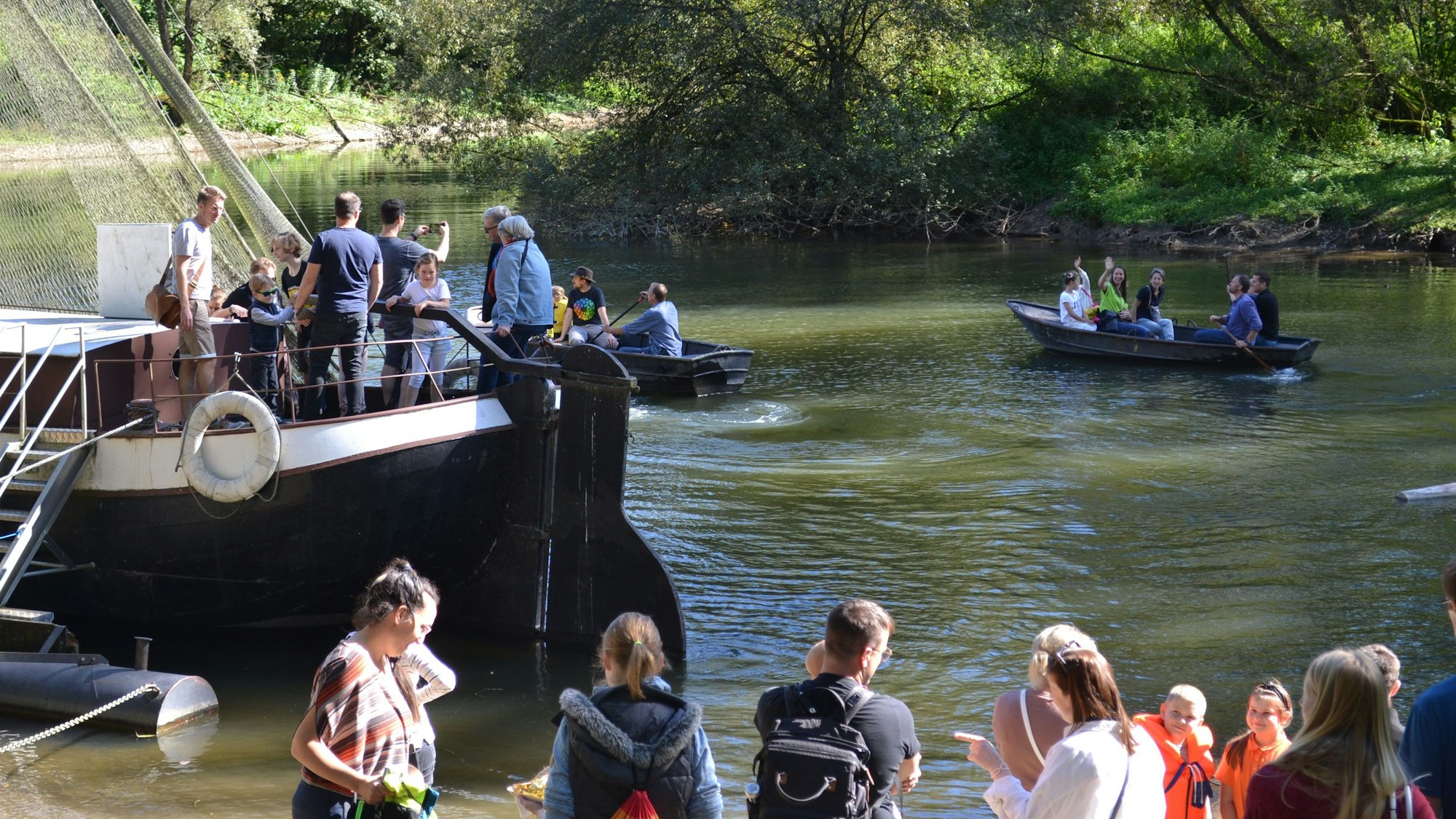 Viele Menschen stehen am Ufer eines Flusses, in Booten und einem großen Aalschokker sind weitere.