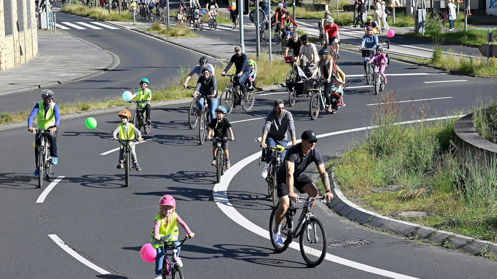 Kinder auf dem Fahrrad mit angebundenen Luftballons sowie Erwachsene fahren um einen Kreisverkehr.