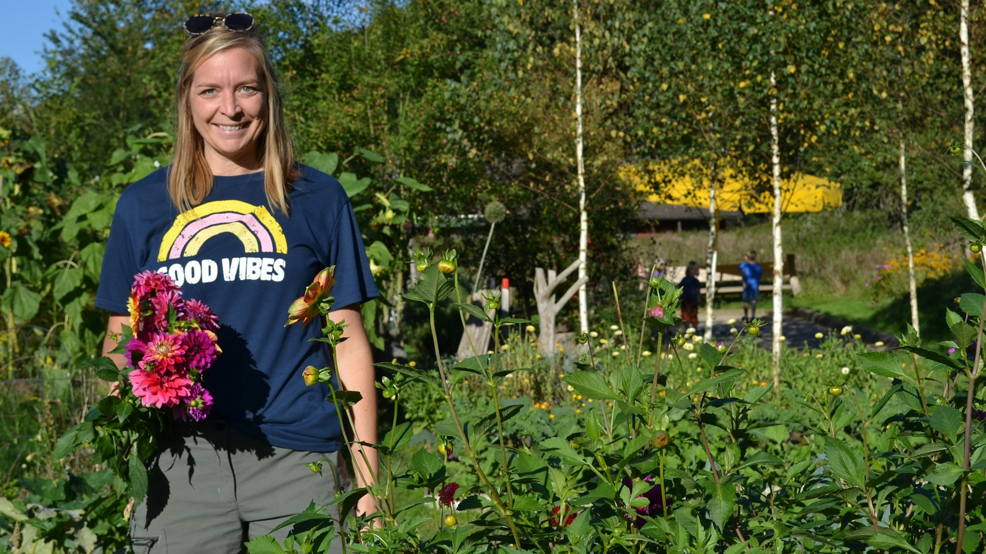 Eine lächelnde Frau mit einem Blumenstrauß aus Dahlien steht ein einem großen, üppigen Garten mit hohen Stauden.