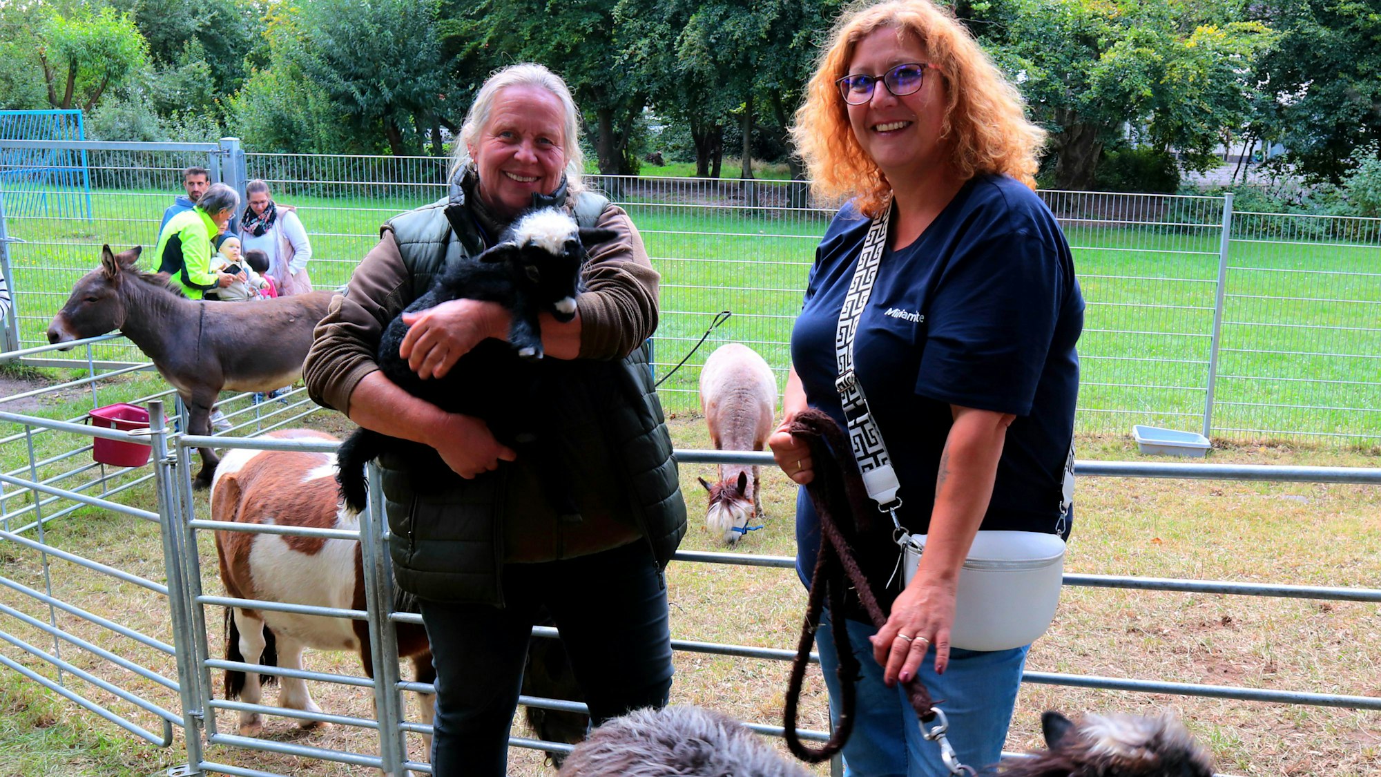 Zwei Frauen stehen vor einem Gehege, in dem ein Esel, ein Pony und ein Alpaka stehen. Eine von ihnen hält ein Lamm auf dem Arm.