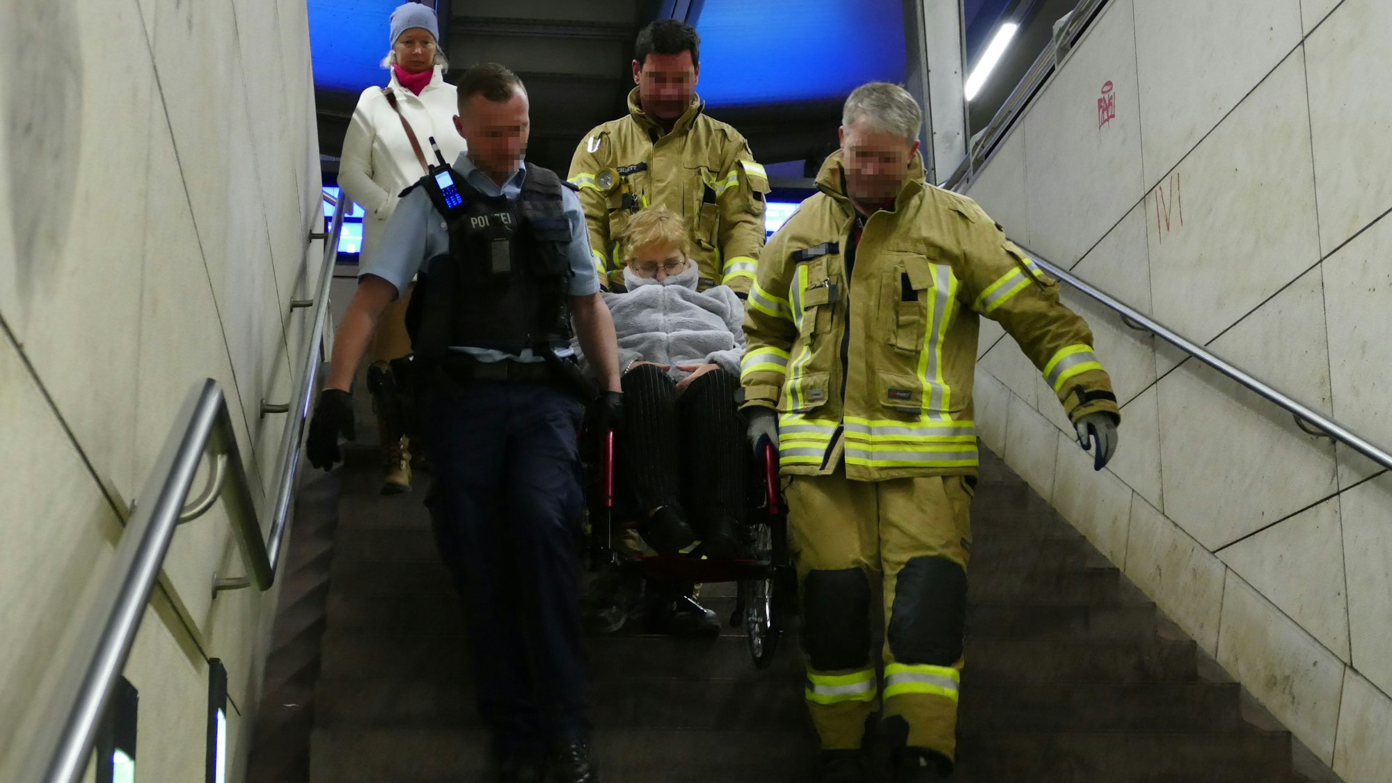 Zwei Feuerwehrmänner in gelben Overalls und ein Bundespolizist in blau tragen eine Frau im Rollstuhl eine Treppe heyrunter.