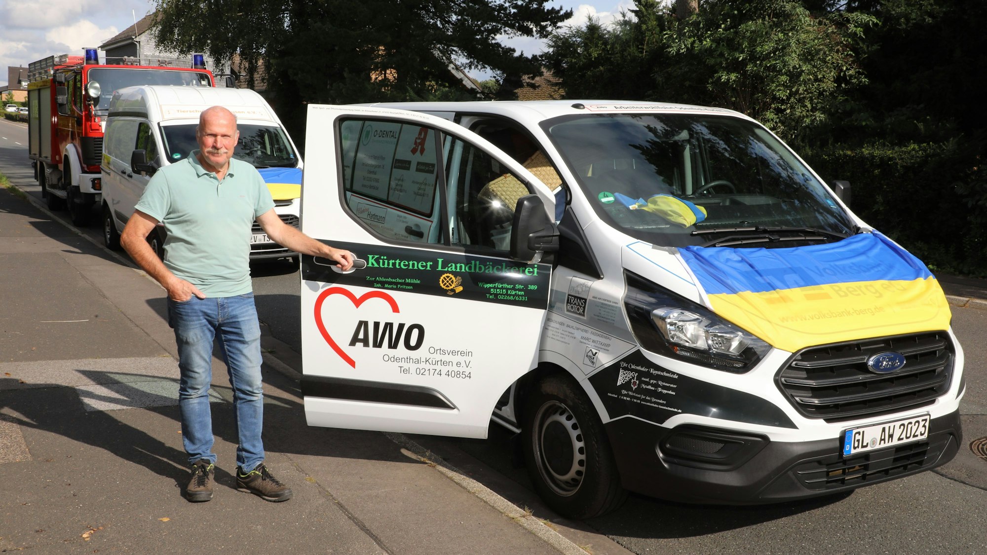 Ein Mann steht an der geöffneten Beifahrertür eines mit einer ukrainischen Fahne geschmückten Transporters. Dahinter steht ein weiterer Transporter und ein Feuerwehrfahrzeug.