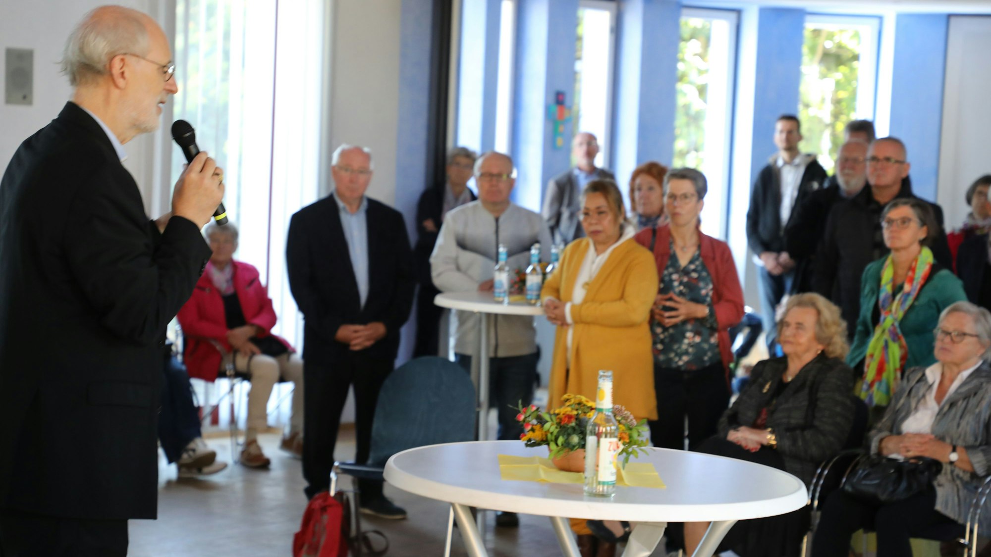 Ein schwarz gekleideter Bischof steht mit einem Mikrofon vor einer Versammlung und spricht.