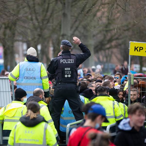 Sicherheitskräfte stehen mit leuchtenden Westen an einem Gitter. Ein Polizist steht auf dem Gitter.
