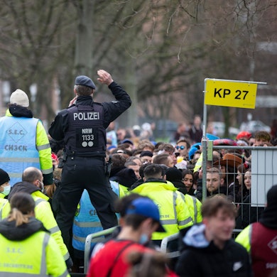 Sicherheitskräfte stehen mit leuchtenden Westen an einem Gitter. Ein Polizist steht auf dem Gitter.