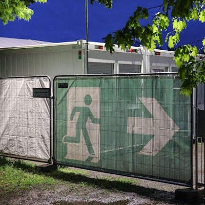 10.05.2023, Nordrhein-Westfalen, Köln: Ein Transparent mit dem Symbol für den Fluchtweg hängt vor Wohncontainer der kommunalen Erstaufnahmeeinrichtung in Köln-Zollstock. Bund und Länder suchen bei einem Spitzentreffen in Berlin nach Lösungen im Streit über die Aufteilung der Kosten für die Versorgung von Flüchtlingen. (Zu dpa: «Gipfel berät über Flüchtlingsfrage») Foto: Oliver Berg/dpa +++ dpa-Bildfunk +++