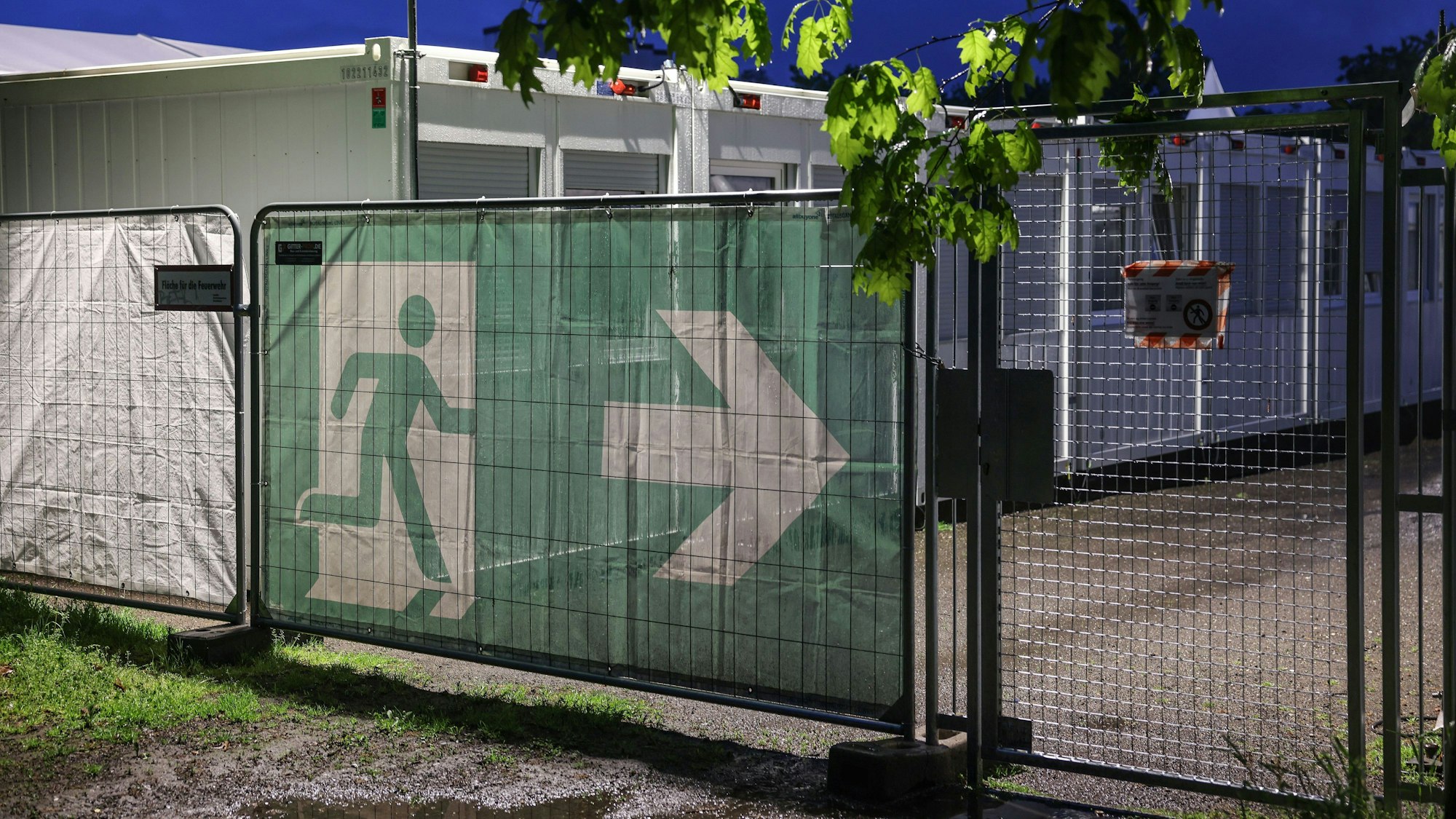 10.05.2023, Nordrhein-Westfalen, Köln: Ein Transparent mit dem Symbol für den Fluchtweg hängt vor Wohncontainer der kommunalen Erstaufnahmeeinrichtung in Köln-Zollstock. Bund und Länder suchen bei einem Spitzentreffen in Berlin nach Lösungen im Streit über die Aufteilung der Kosten für die Versorgung von Flüchtlingen. (Zu dpa: «Gipfel berät über Flüchtlingsfrage») Foto: Oliver Berg/dpa +++ dpa-Bildfunk +++