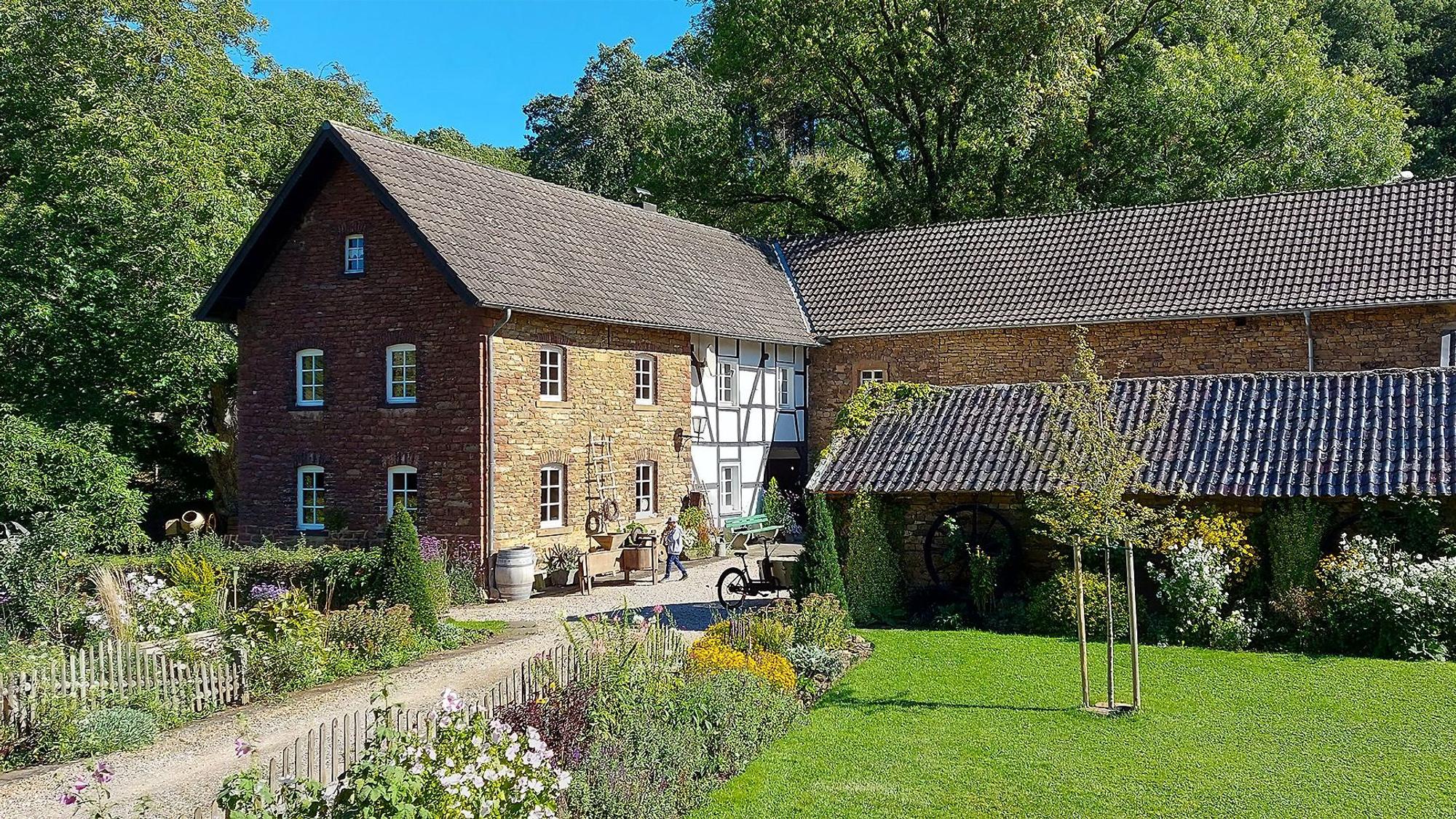 Die Mühle Falkenstein in Eiserfey, umgeben von einem Staudengarten, Wald und einer Wiese.