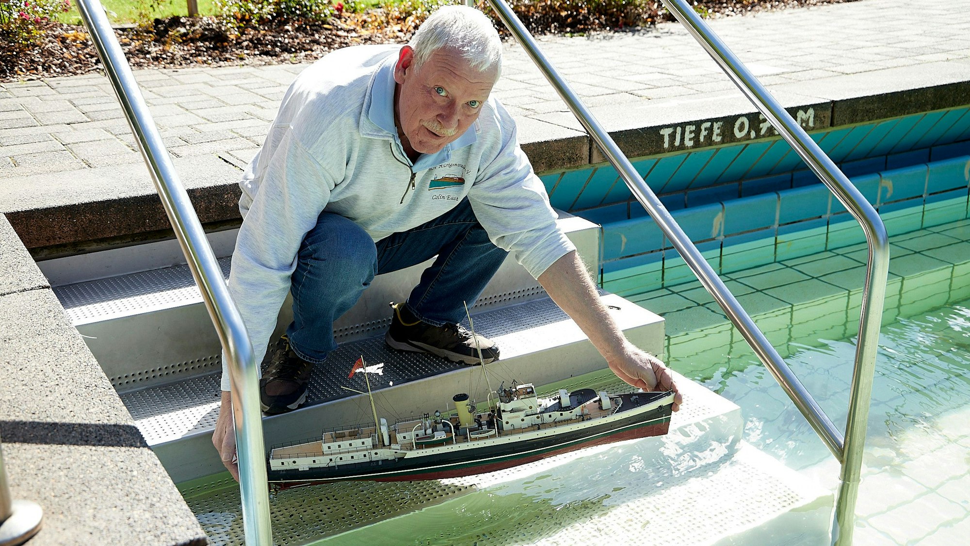 Colin East hockt auf der Treppe, die ins Schwimmbecken des Gemünder Freibads führt. Er hält sein Modellboot in den Händen, das er vorsichtig zu Wasser lässt.