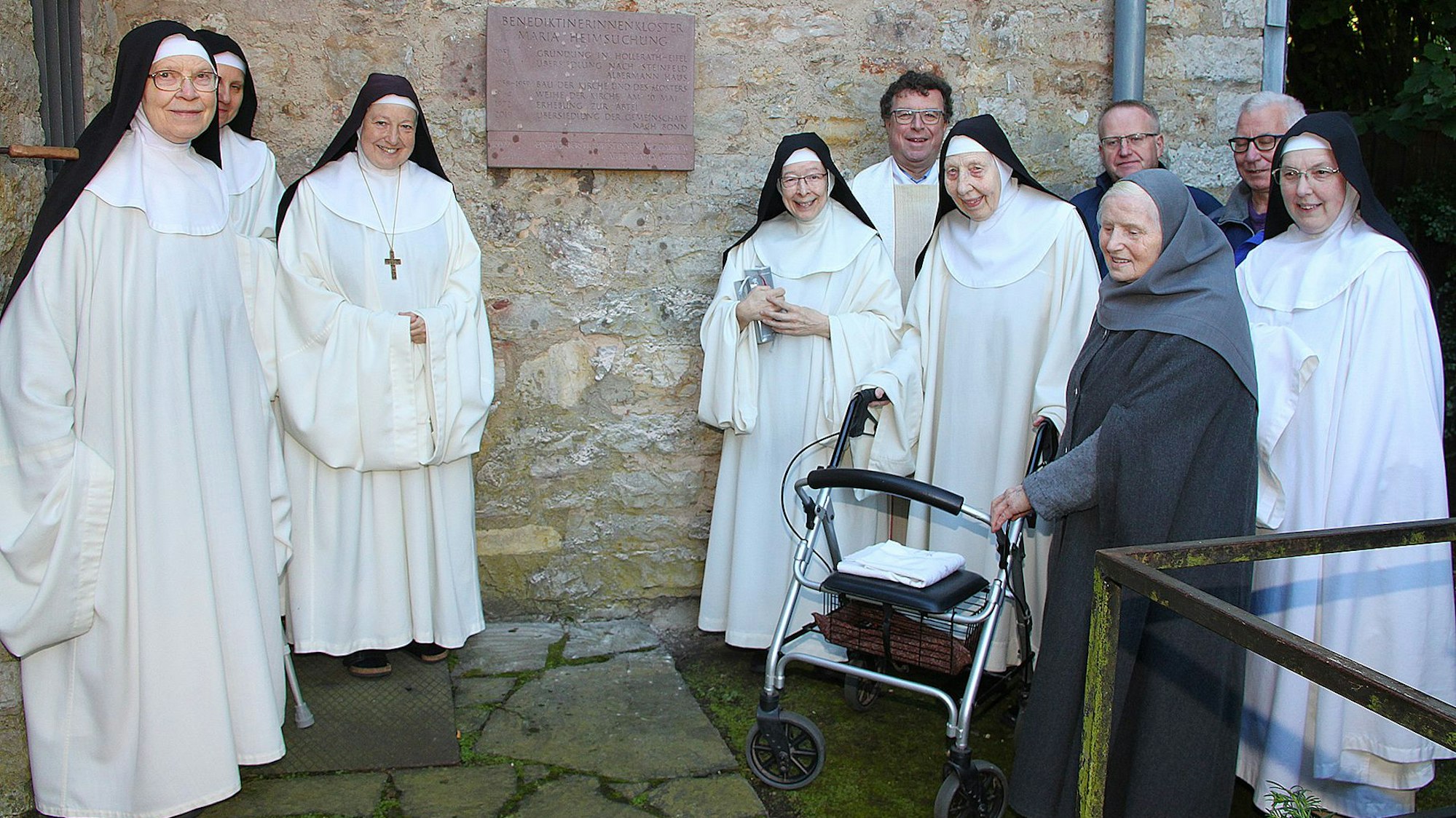 Sechs in Weiß gekleidete Ordensschwestern und eine in Grau gekleidete Eremitin sowie drei Männer im Hintergrund stehen vor einer Tafel, die an einer Mauer angebracht wurde.