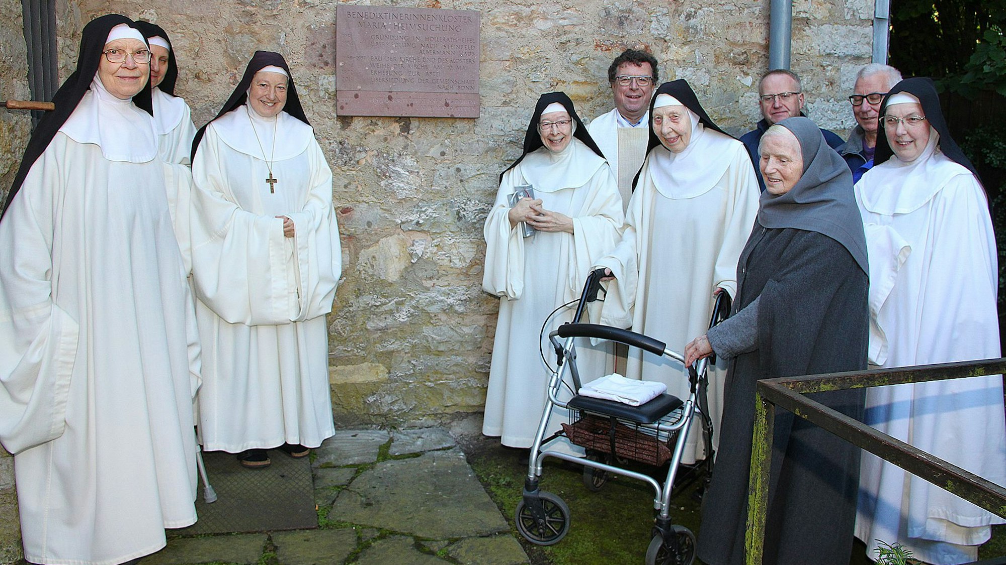 Sechs in Weiß gekleidete Ordensschwestern und eine in Grau gekleidete Eremitin sowie drei Männer im Hintergrund stehen vor einer Tafel, die an einer Mauer angebracht wurde.