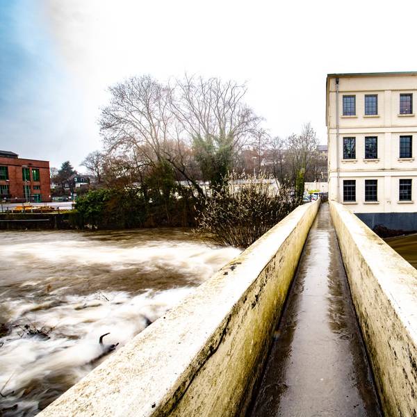 Das Foto zeigt die Fußgängerbrücke über die Wupper mit dem Alten Turbinenhaus und dem Wehr im Hintergrund.