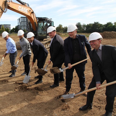 Eine Frau und sechs Männer mit Bauhelm stehen für einen symbolischen Spatenstich neben einem Bagger.