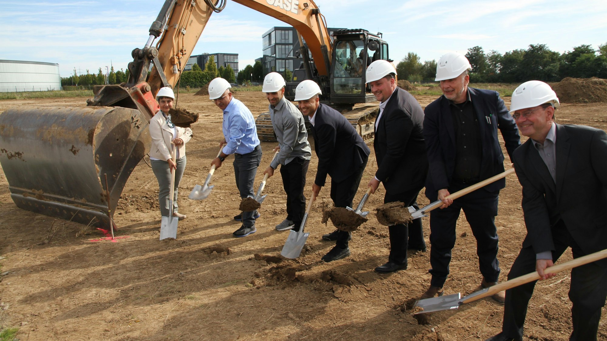 Eine Frau und sechs Männer mit Bauhelm stehen für einen symbolischen Spatenstich neben einem Bagger.