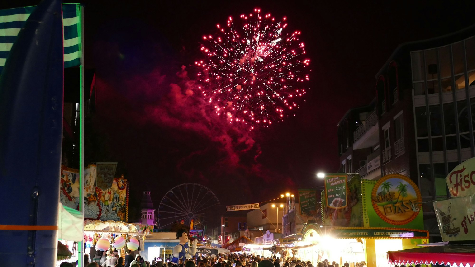 Ein Feuerwerk in Rot strahlt im Nachthimmel über der Kirmes, Menschen stehen dicht gedrängt zwischen den Buden.