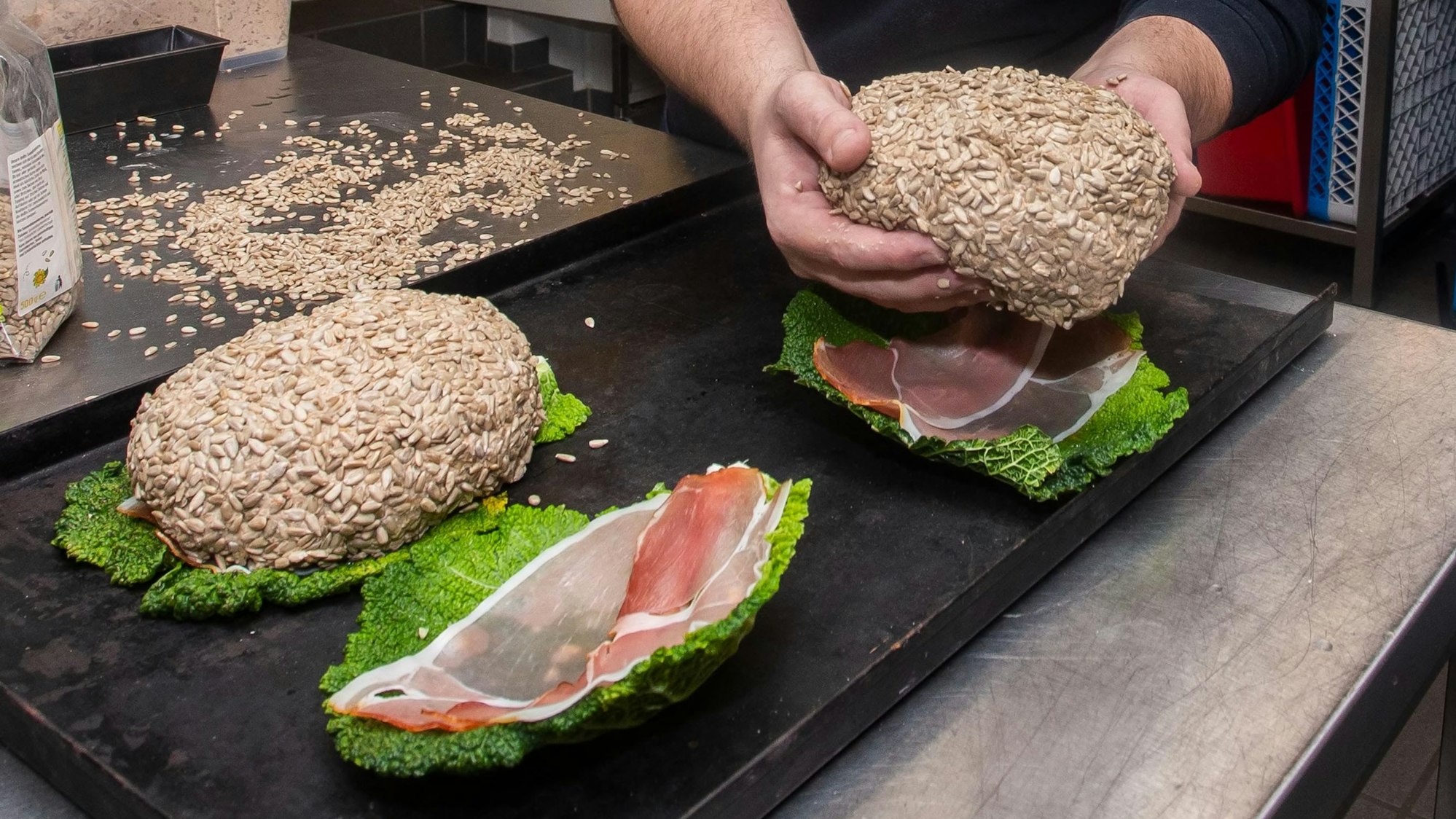 Ein Mann hält ein Brot in der Hand, das er auf ein Bett aus Salatblatt und rohem Schinken legt, bevor es in den Backofen kommt.